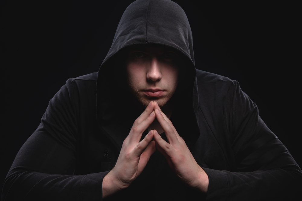 A low-key studio portrait of a large man in a hood sits pondering.