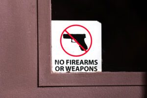 A white square sticker with a hand gun and a no symbol overlaying it and text below that says 'NO FIREARMS OR WEAPONS' in the corner of a door window.